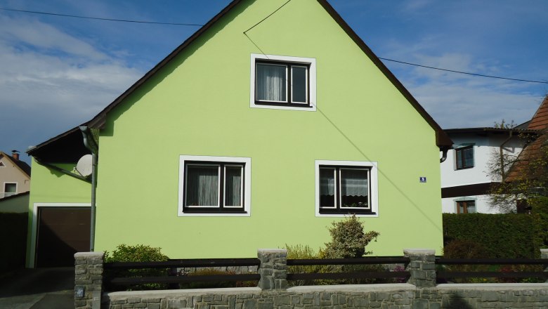 Ein hellgrünes Haus mit Satteldach und zwei Fenstern, umgeben von einem niedrigen Steinzaun.