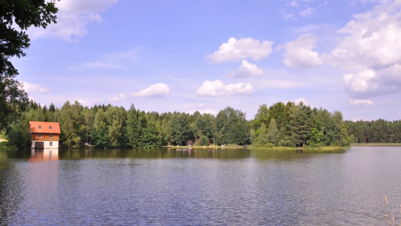 Ein See mit einem Haus am Ufer, umgeben von Wald und blauem Himmel mit Wolken.