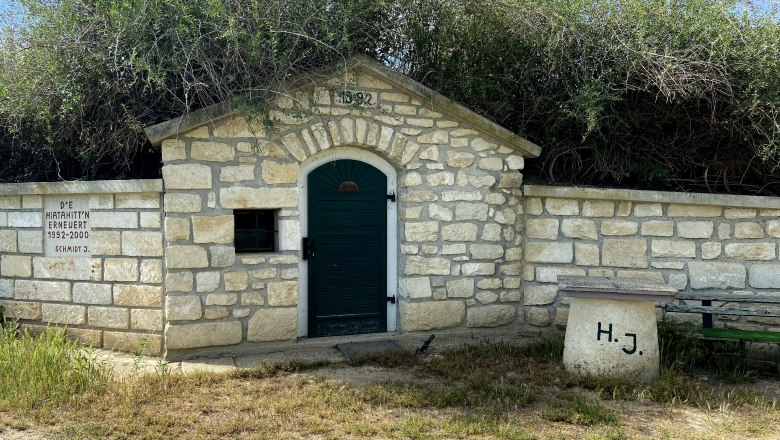 Stone hut with green gate and inscription, surrounded by bushes and grass.