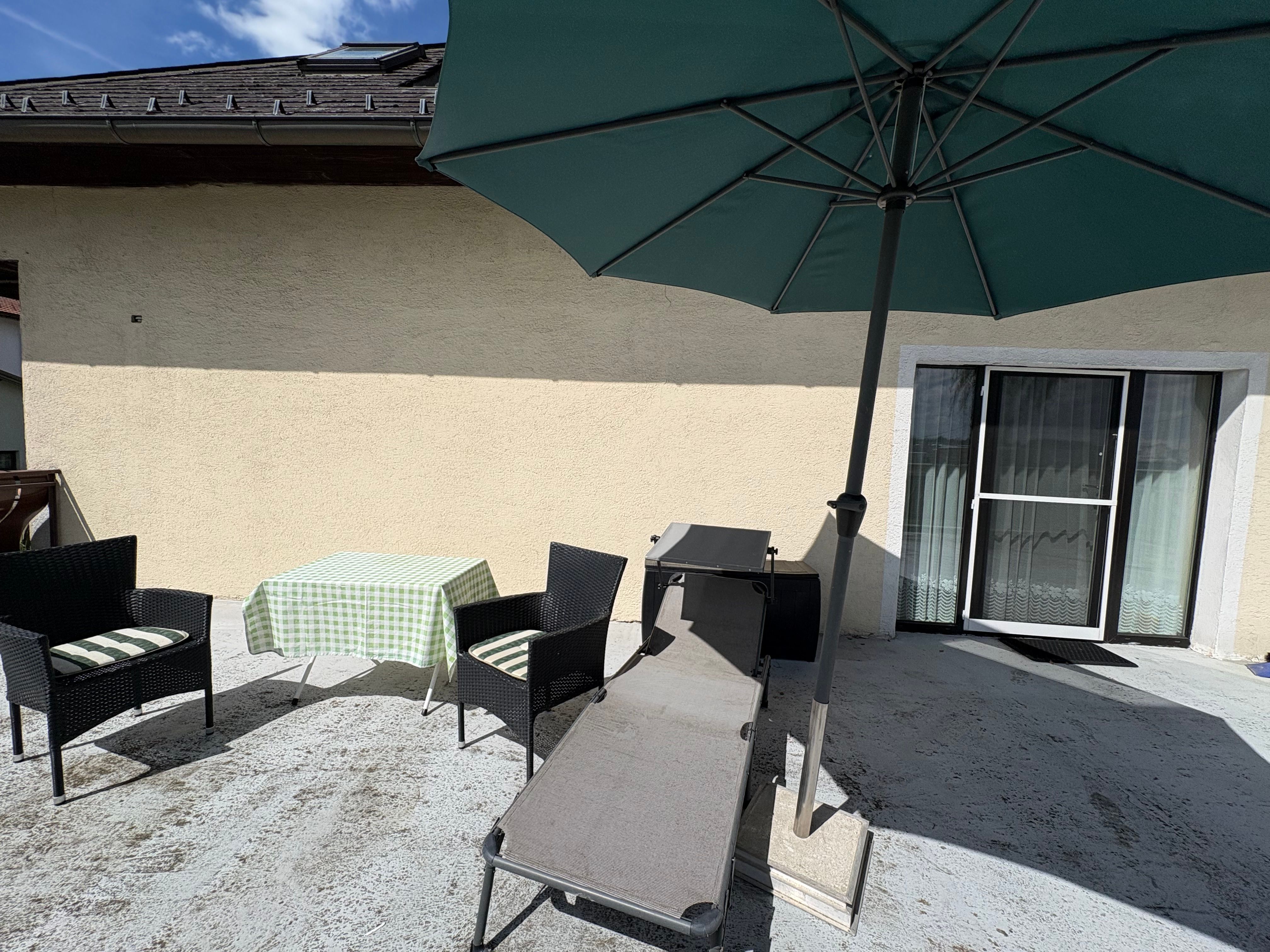 Terrasse mit Tisch, Stühlen, Liege und Sonnenschirm vor einem Haus.