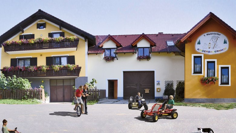 Kinder spielen auf Kettcars vor einem gelben Bauernhof mit Blumenbalkonen.
