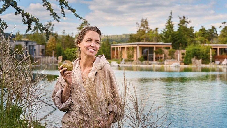 Frau in Bademantel hält Tasse vor einem Teich mit Chalets im Hintergrund.