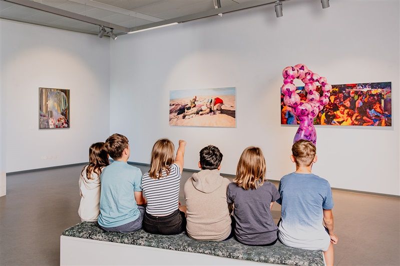 Eine Gruppe von Jugendlichen sitzt auf einer Bank in einem Kunstmuseum und betrachtet Gemälde an der Wand.