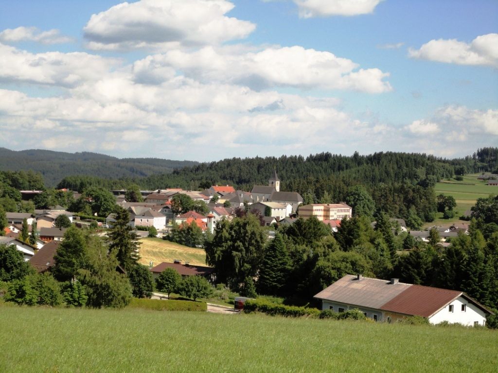 Panorama der Marktgemeinde Martinsberg mit Kirche und umliegenden Häusern, umgeben von Wäldern und Feldern unter blauem Himmel.