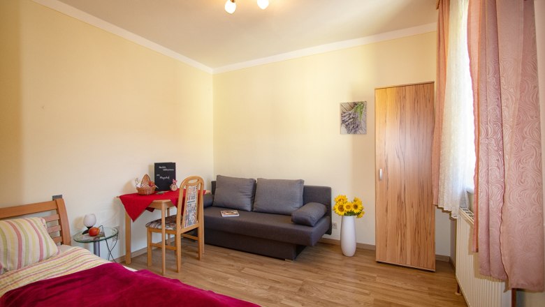 Gemütliches Zimmer mit Bett, Sofa, Tisch und Schrank, dekoriert mit Sonnenblumen und einem Bild an der Wand.