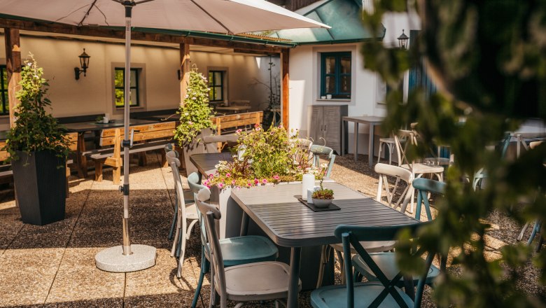Eine sonnige Terrasse mit Tischen, Stühlen und Pflanzen unter einem Sonnenschirm.