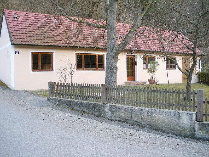 Ein einstöckiges Ferienhaus mit rotem Ziegeldach und hellem Anstrich, umgeben von einem Holzzaun und Bäumen.