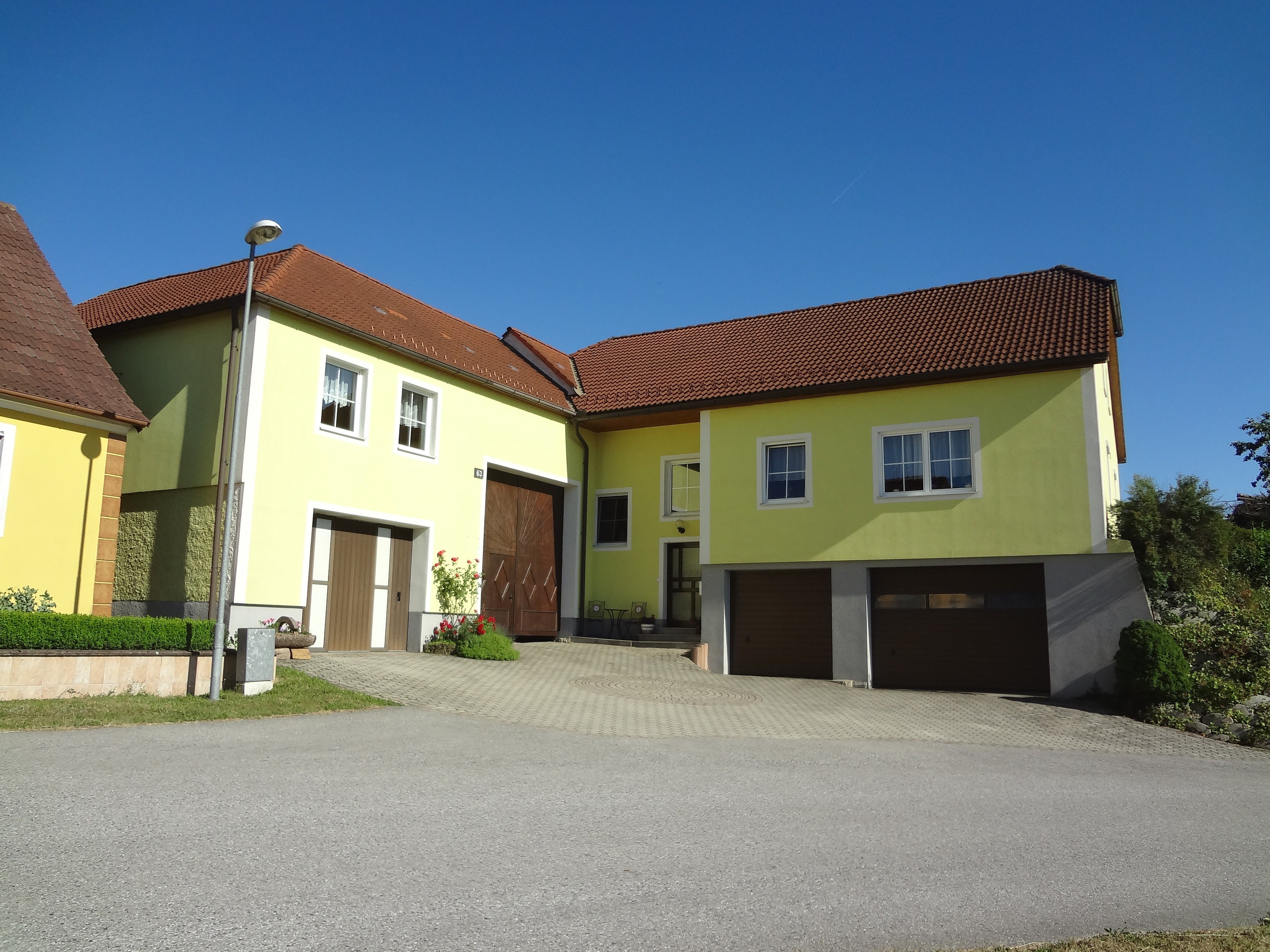 Hausansicht von DSC06383 mit gelber Fassade, roten Ziegeldächern, Doppelgarage und gepflasterter Einfahrt unter blauem Himmel.