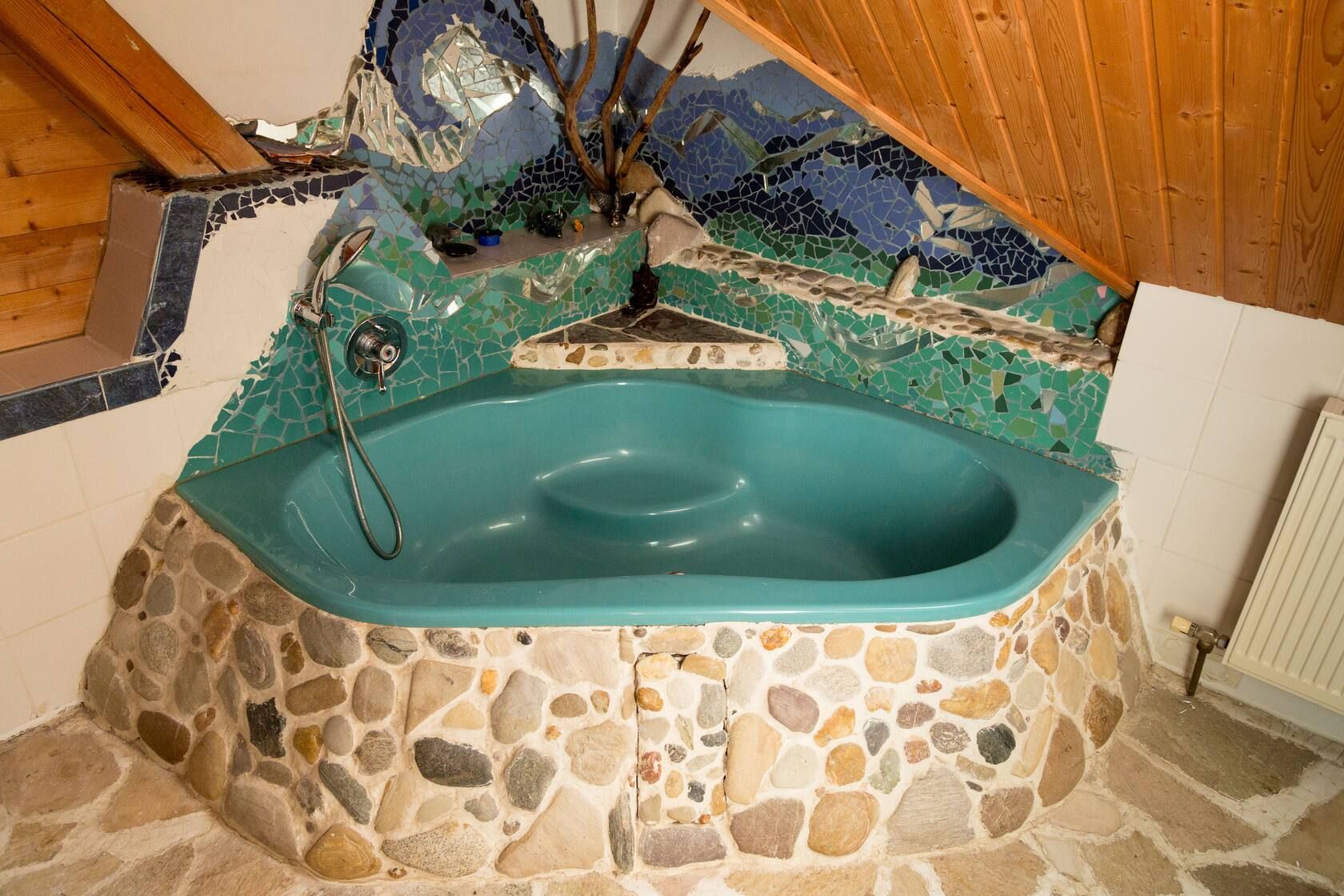 Innenraum mit einer türkisfarbenen Badewanne, Steinverkleidung und Mosaikwand unter einer Holzdecke.