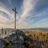 Ein Gipfelkreuz auf einem Felsen mit einer Aussichtsplattform, umgeben von Wald und einem weiten Blick über die Landschaft unter einem blauen Himmel.