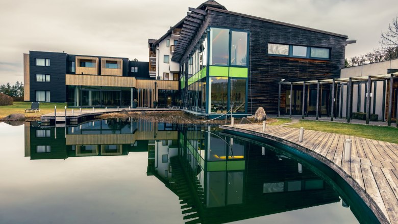 Modernes Wellnesshotel mit Holzfassade und großem Teich im Vordergrund.