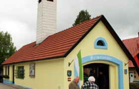 Kleines gelbes Gebäude mit rotem Dach und Schornstein, beschriftet mit 'Heimatstube'. Zwei Personen stehen davor.