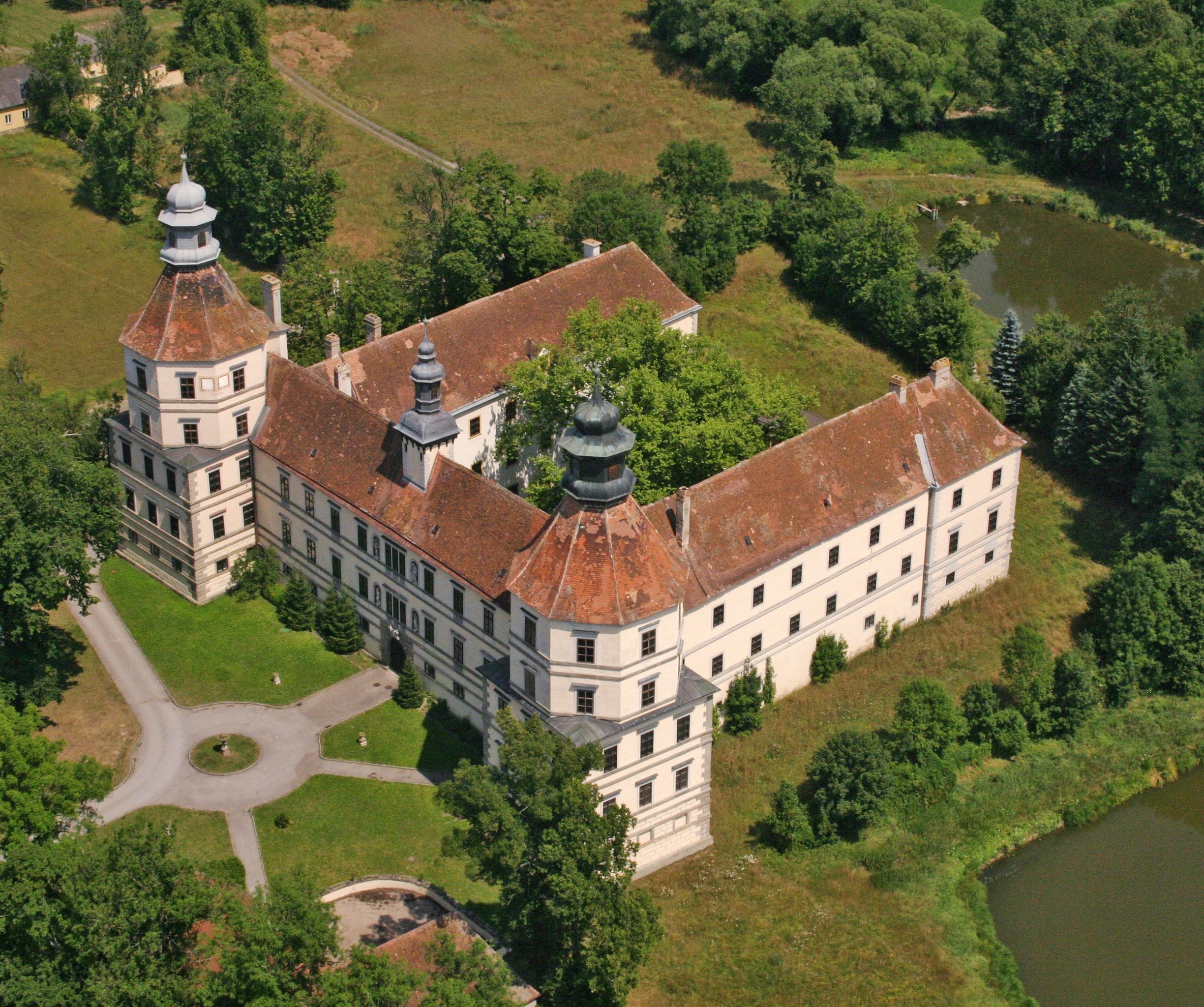 Luftaufnahme eines historischen Schlosses mit zwei Türmen und einem Innenhof, umgeben von Bäumen und einem Teich.