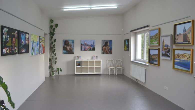 Innenansicht einer Galerie mit Gemälden an den Wänden und einem Regal in der Mitte.