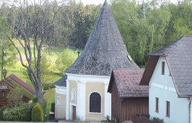 Eine kleine Kapelle mit spitzem Dach, umgeben von Bäumen und Häusern.