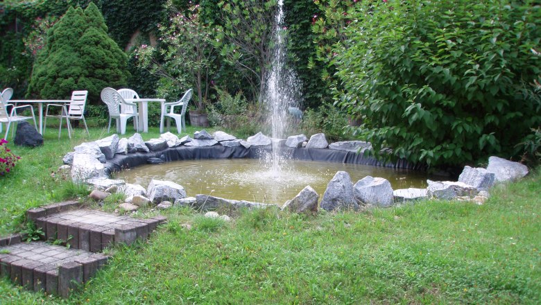 Garten mit kleinem Teich und Springbrunnen, umgeben von Pflanzen und Gartenmöbeln.