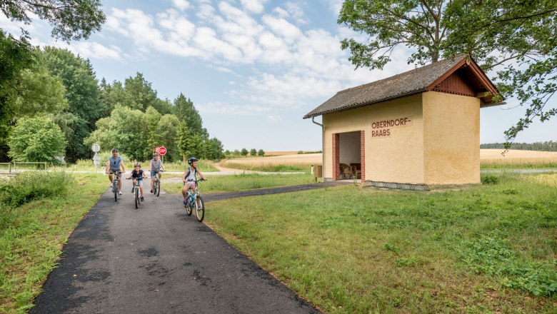 Eine Familie fährt mit Fahrrädern auf einem Weg neben einem gelben Gebäude mit der Aufschrift 'Oberndorf-Raabs'.