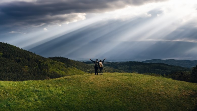 Zwei Personen stehen auf einem Hügel mit Rucksäcken, umgeben von grünen Wäldern und dramatischen Sonnenstrahlen durch Wolken.