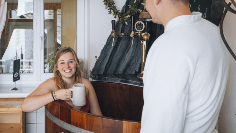 Frau in einem Bierbad mit Krug, Mann im Bademantel daneben.