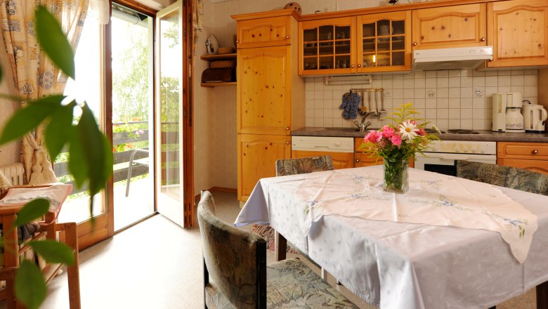 Gemütliche Küche mit Holzmöbeln, Esstisch und Blumenstrauß. Offene Balkontür mit Blick ins Grüne.