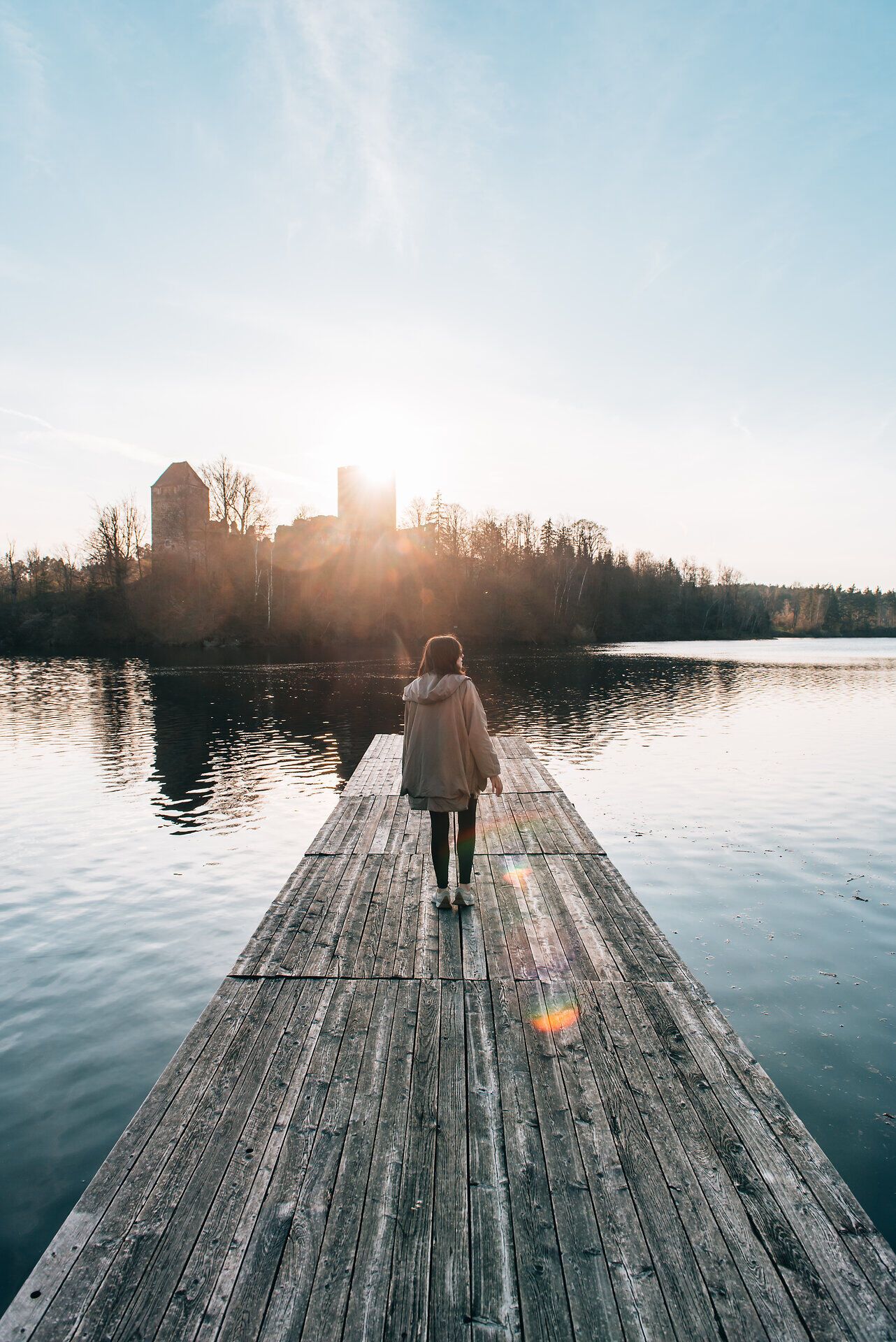 Ein sanfter Sonnenuntergang taucht die Landschaft in warmes Licht, während eine Person auf einem Holzsteg dem ruhigen Wasser entgegengeht. Die Stille der Natur und die sanften Wellen schaffen eine friedliche Atmosphäre, die zum Verweilen einlädt.