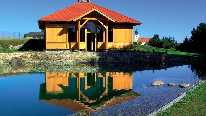 A wooden building with a red roof is reflected in a pond surrounded by a green landscape.
