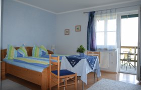 Gemütliches Gästezimmer mit Doppelbett, Tisch und Stühlen, blauen Vorhängen und Zugang zu einem Balkon mit Sitzgelegenheit.