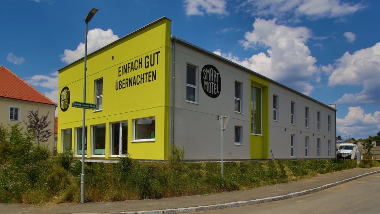 Smart Motel Außenansicht, © Smart Motel GmbH Außenansicht eines modernen, zweistöckigen Motels mit gelber Fassade und der Aufschrift 'Einfach gut übernachten'.