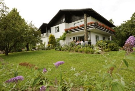 Ein zweistöckiges weißes Haus mit Balkon, umgeben von einem grünen Garten mit Blumen und Bäumen.
