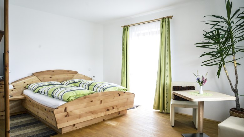 Helles Zimmer mit Holzbett, grün-weiß gestreifter Bettwäsche, Tisch mit Vase und Pflanze, grüne Vorhänge.