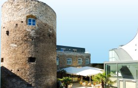 Historischer Turm neben modernem Gebäude mit Terrasse und Sonnenschirmen.