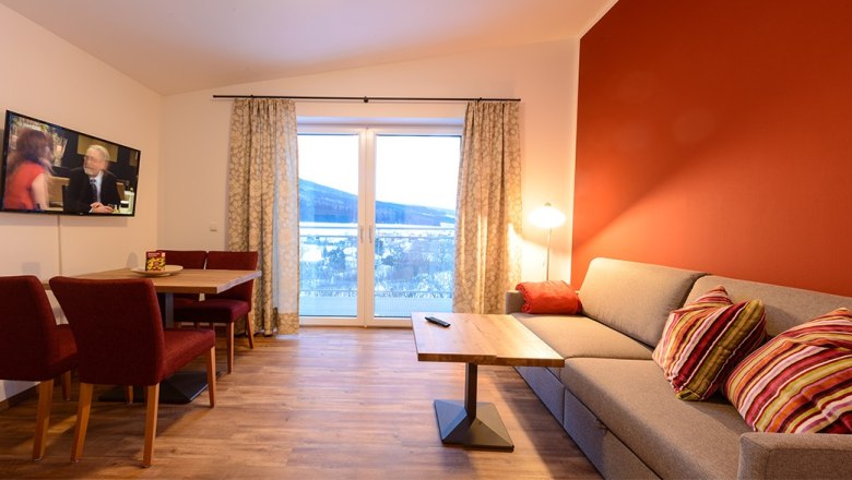 Cozy living room with red accent wall, grey sofa, wooden table, TV and large window with curtains.