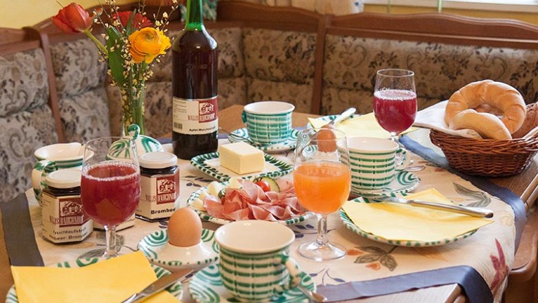Ein gedeckter Frühstückstisch mit Brötchen, Marmelade, Eiern, Saft und Blumen.