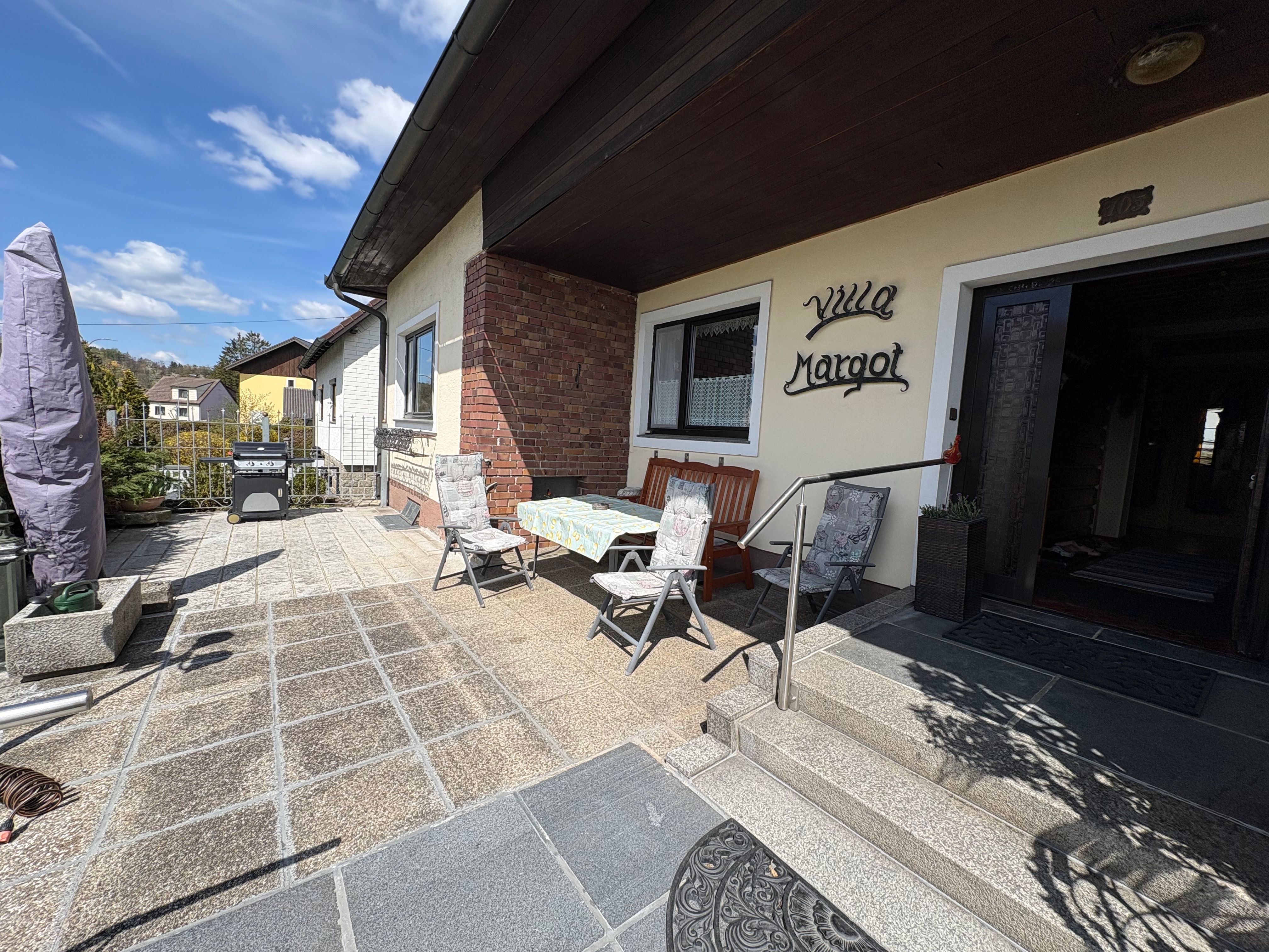 Eingang einer Villa mit Terrasse, Tisch und Stühlen, Grill und Pflanzen.
