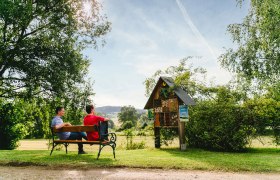 Zwei Personen sitzen auf einer Bank im Grünen vor einem Insektenhotel.