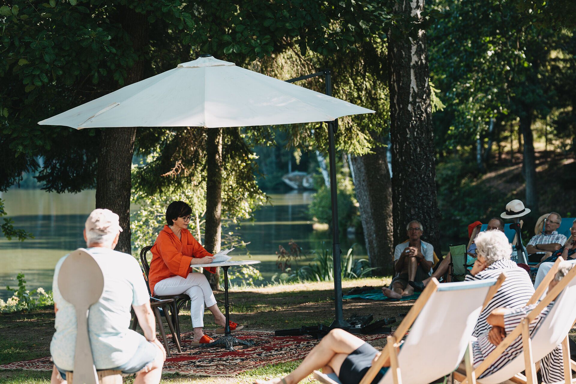 Unter einem schattenspendenden Sonnenschirm versammeln sich die Zuschauer, um den Darbietungen des Theaterfestivals HIN & WEG zu lauschen. Die sanfte Brise und das Plätschern des Wassers im Hintergrund schaffen eine entspannte Atmosphäre, die den kulturellen Genuss im Waldviertel perfekt ergänzt.