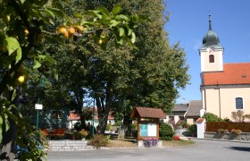 Dorfplatz mit Kirche, Bäumen und Infotafel in Eggern.