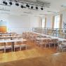 Ein leerer Saal mit Holzboden, Tischen und Stühlen, einer Bühne und Beleuchtung an der Decke.