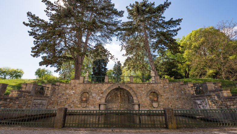 Steinmauer mit zwei großen Bäumen und dekorativen Elementen im Schlossgarten.