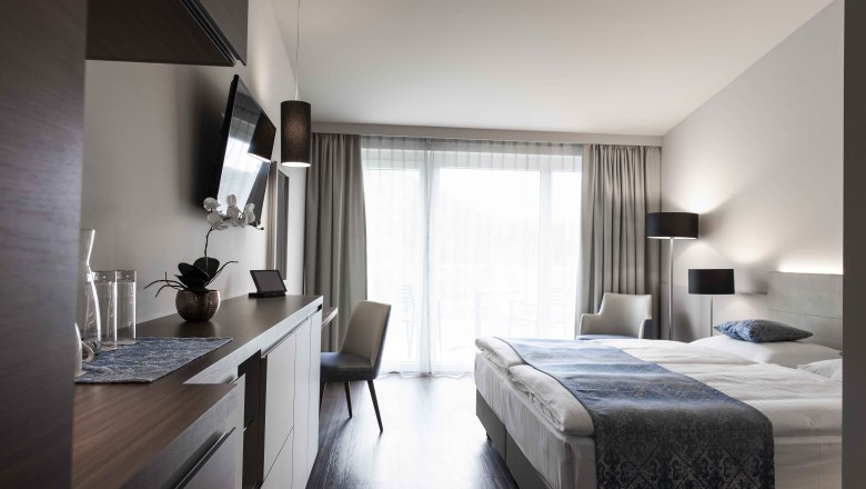 Modernes Hotelzimmer mit Doppelbett, Fernseher, Schreibtisch und großen Fenstern mit Vorhängen.