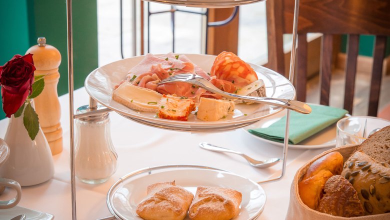 Ein Frühstückstisch mit einer Etagere voller Aufschnitt, Marmeladen und Brötchen, daneben ein Glas Orangensaft.
