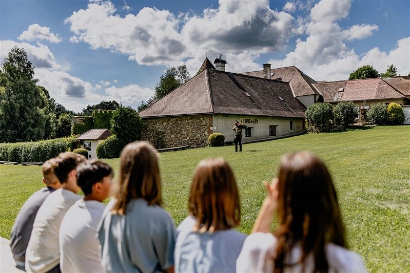 Eine Gruppe von Jugendlichen sitzt auf einer Wiese und schaut auf ein Gebäude, vor dem eine Person steht. Der Himmel ist bewölkt.