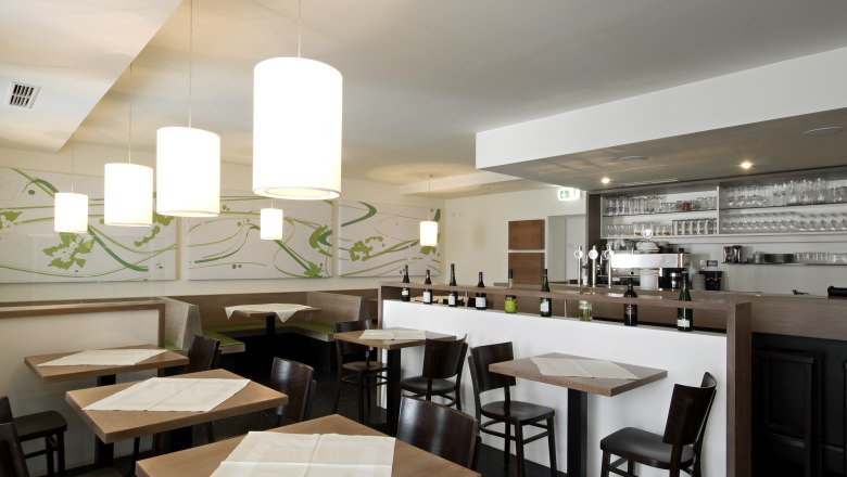 Innenansicht eines modernen Restaurants mit Holztischen, Stühlen und einer Bar mit Flaschen und Gläsern.