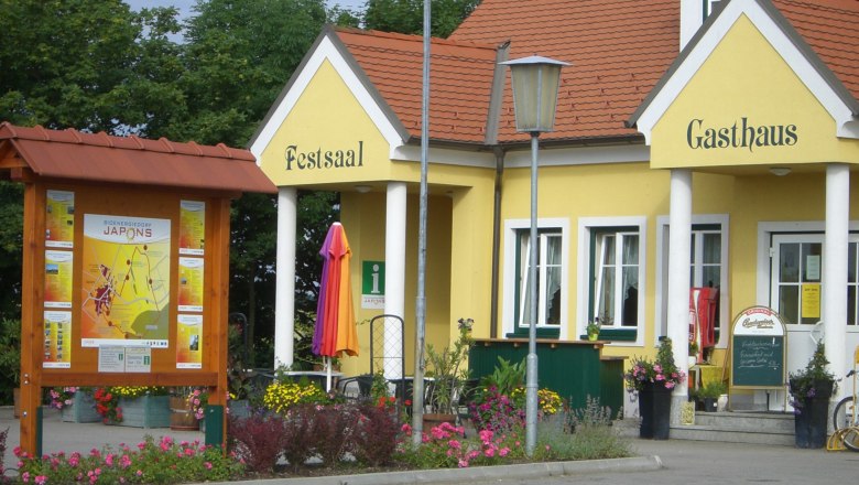 Gelbes Gebäude mit der Aufschrift 'Festsaal' und 'Gasthaus', umgeben von Blumen und einem Informationsschild.