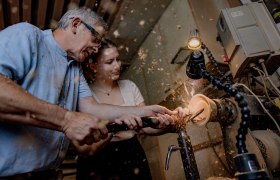 Zwei Personen arbeiten gemeinsam an einer Drehbank, um ein Holzstück zu bearbeiten. Holzspäne fliegen in der Luft.