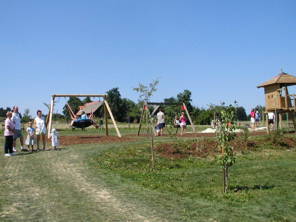 Ein Spielplatz mit Schaukeln, Klettergerüst und Kindern, die spielen. Erwachsene stehen daneben. Es ist ein sonniger Tag.