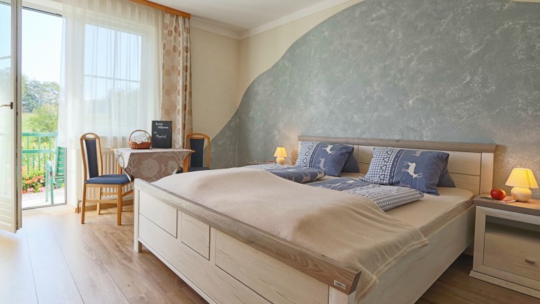 Gem&uuml;tliches Schlafzimmer mit Doppelbett, blauer Bettw&auml;sche, Tisch mit St&uuml;hlen und Balkonzugang. Helle, einladende Atmosph&auml;re.