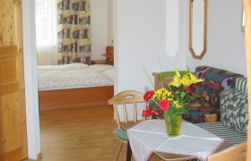 Ein gemütliches Zimmer mit Holzmöbeln, einem Bett im Hintergrund und einem Tisch mit Blumen im Vordergrund.