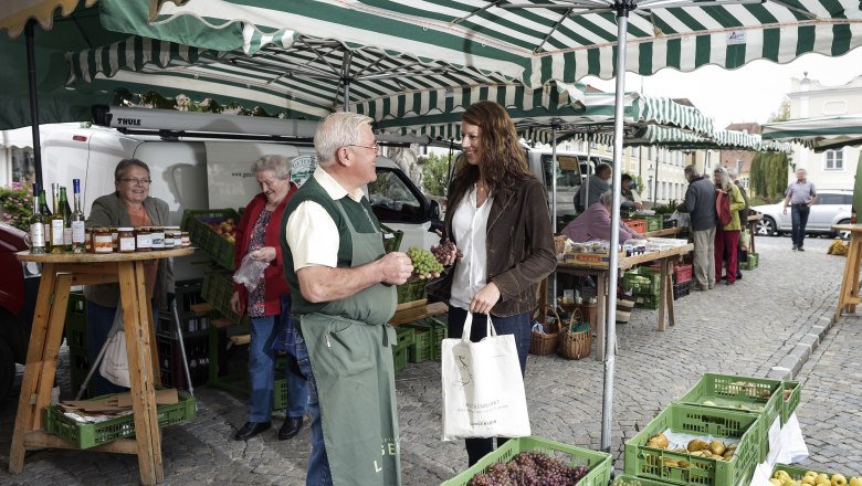 Menschen auf einem Wochenmarkt mit Obst- und Gemüseständen unter gestreiften Markisen.