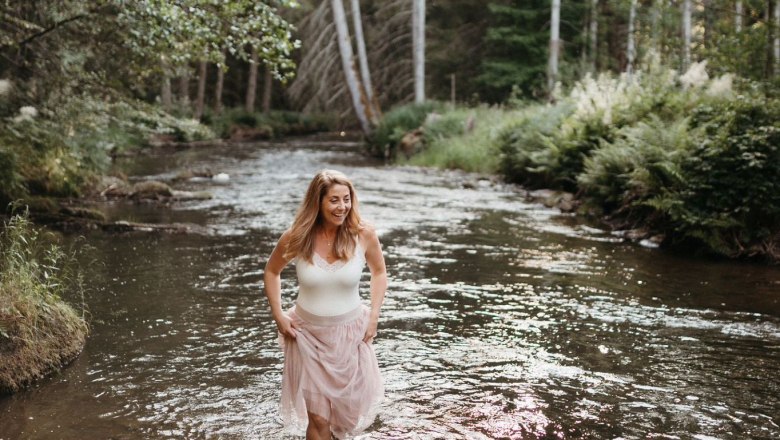 Eine Frau steht lächelnd in einem flachen Fluss, umgeben von einem Wald bei Sonnenuntergang.